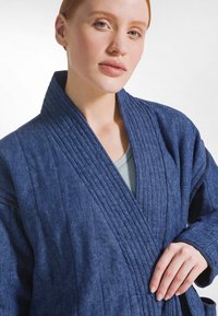 Námořnicky modrý župan z texturované látky s hlubokým límcem, vertikálním žebrováním a volným střihem, nošený přes světlý top.