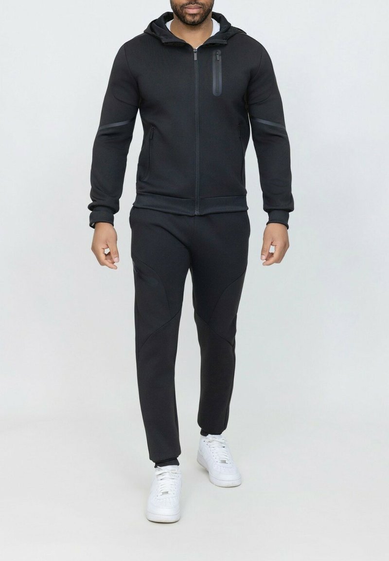 Haut à capuche noir zippé avec une coupe slim, doté d'une poche sur la poitrine, associé à un pantalon noir fuselé. Des baskets blanches complètent la tenue.