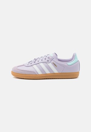 Lawendowy trampek Adidas Samba z białymi paskami, gumową podeszwą, miętową zakładką na pięcie i złotym tekstem "Samba" z boku.