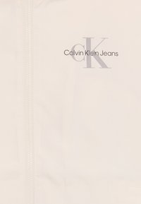 Lys beige jakke med frontglidelås, glatt tekstur og trykt logo: "Calvin Klein Jeans" med en stilisert 'CK' i grått.