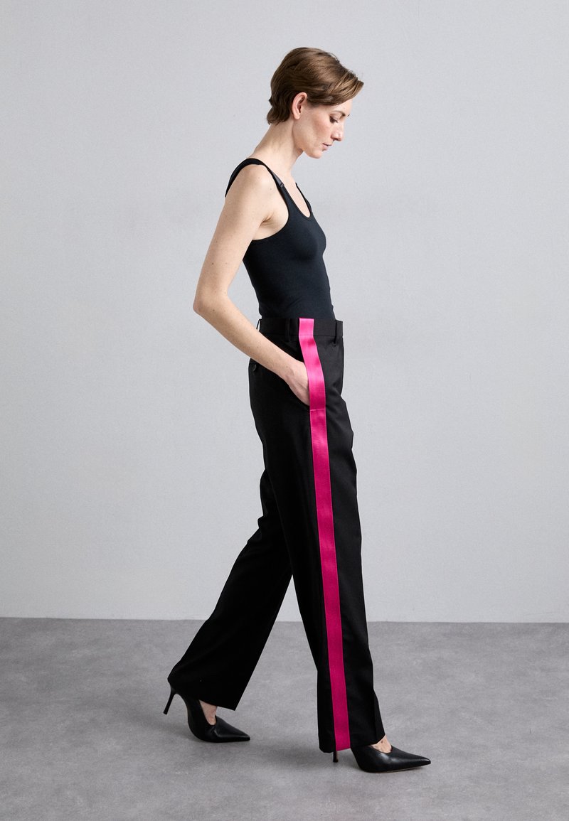 Pantaloni neri con una striscia laterale rosa brillante, caratterizzati da un design a gamba dritta e tasche, abbinati a una canotta nera e tacchi a spillo neri.