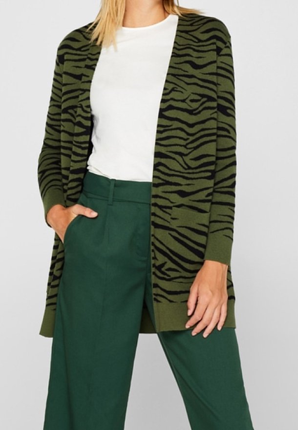Femme portant un cardigan à imprimé zèbre vert et noir, un haut blanc et un pantalon taille haute vert foncé, la main dans la poche, debout devant un fond gris.