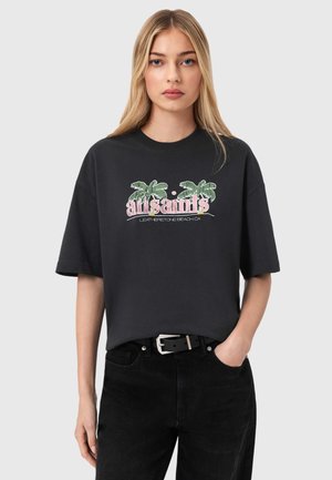 Ung kvinde med langt blondt hår iført en sort oversized t-shirt med palmetræsgrafik og teksten "all saints", kombineret med sorte jeans og bælte.