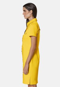 Gelbes Kurzarmkleid mit durchgehender Knopfleiste, zwei Brusttaschen und Seitenschlitzen. Aus leichtem Stoff mit gerader Silhouette.