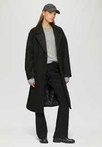 Long manteau noir texturé avec des revers larges, pull en maille gris et pantalons noirs évasés. Chaussures noires cloutées. Une casquette grise complète la tenue.
