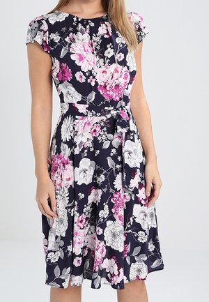 Nainen, joka pukeutuu polvipituiseen laivastonsiniseen mekkoon, jossa on valkoisia ja vaaleanpunaisia kukka- ja lintukuoseja, lyhyet hihat ja samaa kangasta oleva vyö solmittuna vyötärölle.