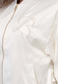 Witte satijnen bomberjack met een gouden rits, ribbelkraag en geborduurd logo op de borst. Inclusief zijzakken.