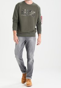 Felpa verde oliva con stampa mimetica 'ALPHA INDUSTRIES'. Abbinata a jeans grigi sbiaditi e stivali stringati color sabbia. Design semplice, vestibilità casual.