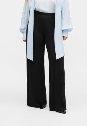 Άτομο που φορά ψηλόμεσο, φαρδύ παντελόνι σε μαύρο χρώμα, ανοιχτό μπλε μπλούζα με μακριά μανίκια και εκτεταμένα μανσέτα, καθώς και μαύρα στενά παπούτσια, όρθιο.