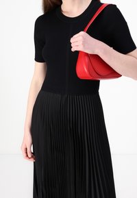 Žena v černém žebrovaném topu a plisované sukni, držící malou červenou koženou kabelku přes rameno na bílém pozadí.
