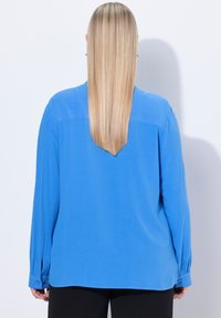 Blusa blu a maniche lunghe con una superficie liscia, polsini abbottonati e una linea dritta. Cuciture visibili e dettaglio a tramatura sulla parte posteriore.