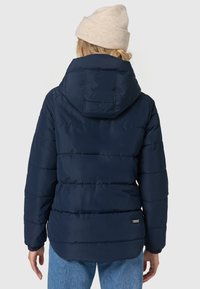Marineblå pufferjakke med vandrette syninger og hætte, fremstillet af skinnende vandafvisende materiale. Etiketten er synlig nederst til venstre.