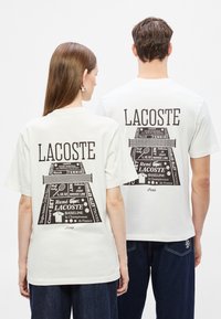 Λευκά βαμβακερά T-shirts με μαύρο γραφικό εκτύπωμα στοιχείων γηπέδου τένις και κείμενο "LACOSTE" στην πλάτη. Κοντά μανίκια, άνετη εφαρμογή.