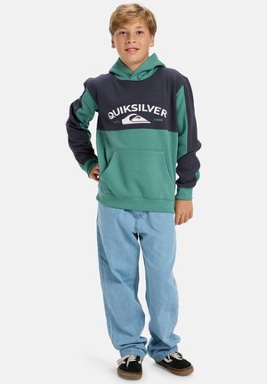 Green and navy hoodie with a front pocket and white logo, paired with light blue jeans and black shoes with white laces.
