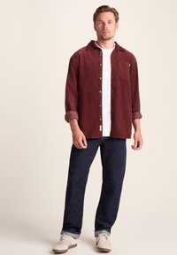 Maroon button-up shirt with a pointed collar, unbuttoned at the top, paired with dark blue jeans and light sneakers.