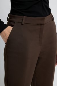 Persoon in een paarse nauwsluitende broek met een hand in de zijzak en een zwart langemouwen top zichtbaar bij de taille en pols.