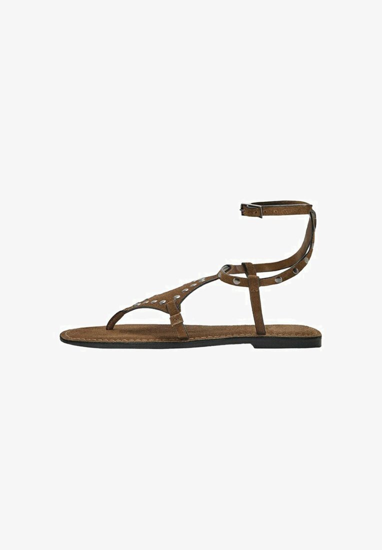 Brun flad sandal med ankelrem og sølvnitter på tårem og sidelommer, med sort sål og syninger langs kanterne.