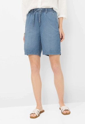 Vrouw draagt blauwe denim shorts met fijne strepen, een elastische tailleband en een trekkoord, gecombineerd met witte sandalen en een wit bloesje.