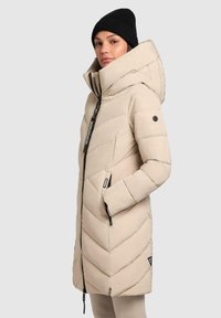 Beige pufferjack met een hoge kraag, zigzag stiksel en ritssluiting. Voorzien van een logo op de mouw en voorzakken, gecombineerd met een zwarte gebreide muts.