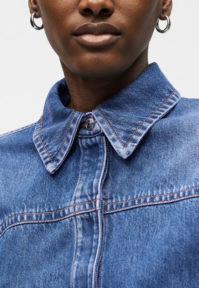 Κοντινό πλάνο ενός ατόμου που φοράει μπλε τζιν μπουφάν με κούμπωμα στον γιακά, με ασημένιους κρίκους σκουλαρίκια ορατούς και στα δύο αυτιά.