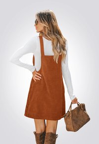 Braunes Cordkleid mit Trägern, knielang, über einem weißen Langarmshirt getragen. Das Modell hält eine braune Handtasche und trägt kniehohe Stiefel.