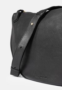 Black leather bag with a pebbled texture, featuring a wide adjustable strap and a gold accent on the strap. Engraved logo on the front.