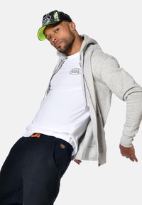 Hoodie gris zippé superposé à un t-shirt blanc avec un logo, associé à un pantalon de jogging noir avec une étiquette orange. Casquette noire et verte.