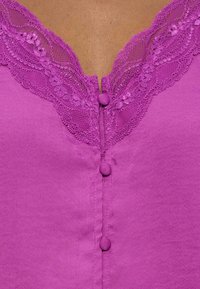 Μωβ μπλουζάκι χωρίς μανίκια με δαντελένιο τελείωμα και κουμπιά καλυμμένα με ύφασμα κατά μήκος του μπροστινού λαιμού σε μέτριο καφέ δέρμα.