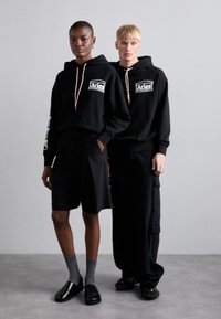 Twee modellen dragen zwarte hoodies met logo's en contrasterende trekkoorden. Het ene paar is gestyled met zwarte shorts, het andere met cargobroeken en schoenen.