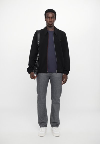 Veste zippée noire avec un col structuré, t-shirt bleu marine à col rond, jeans gris droit et baskets blanches. Sac à bandoulière noir.