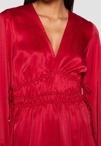 Robe en satin rouge avec un décolleté profond en V, manches longues, taille élastique froncée et liens latéraux portée par une personne du cou jusqu'à la taille.