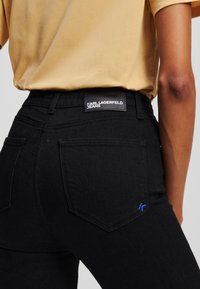 Jeans de mezclilla negra de cintura alta, con un parche de logo en la parte posterior y costuras azules sutiles en los bolsillos traseros. Textura suave.
