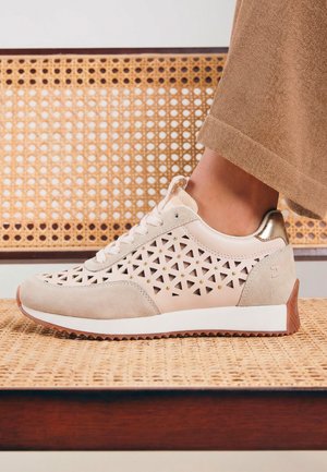 Baskets beiges avec découpes triangulaires, clous dorés, semelle blanche et semelle extérieure marron, portées avec un pantalon marron clair sur un siège de chaise en osier.