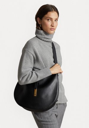 Black leather shoulder bag with a curved shape, featuring a gold accent and a long strap, held by a model in a gray turtleneck sweater.