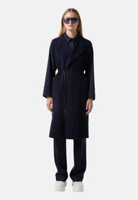 Navy wool blend trench coat with side pockets, a belted waist, and lapel collar; worn over dark trousers and white platform sneakers.