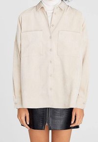 Camicia beige con bottoni realizzata in tessuto morbido, dotata di due tasche frontali e una vestibilità comoda, abbinata a una gonna in pelle nera texture.