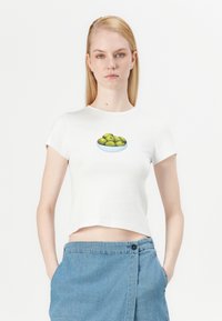 Camiseta recortada blanca con un gráfico de aceitunas verdes en un tazón en el pecho, combinada con pantalones cortos de mezclilla azul claro.