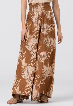 Pantalon marron à jambes larges avec imprimé de feuilles de palmier beiges et taille élastique froncée, porté avec des sandales tressées sur fond clair.