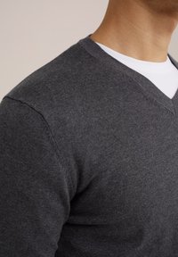 Grau V-Ausschnitt-Pullover aus weichem Stoff, mit figurbetontem Design und glatter Textur, über einem weißen Rundhals-Shirt getragen.