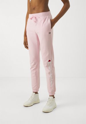 Persona que lleva pantalones deportivos Champion de color rosa claro y zapatillas blancas, de pie con una mano en el bolsillo contra un fondo blanco liso.