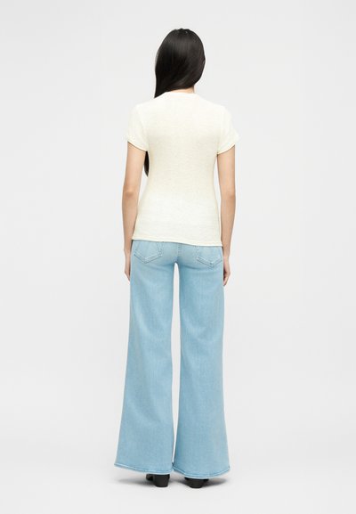 Γυναίκα με μακριά σκούρα μαλλιά που φοράει κοντομάνικη κρεμ μπλούζα και ανοιχτό μπλε ψηλόμεσα παντελόνια με φαρδιά πόδια, στέκεται με την πλάτη προς το μέρος μας σε λευκό φόντο.