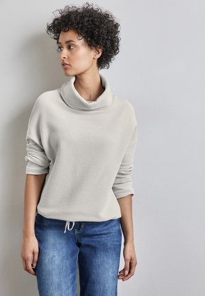 Pull gris clair texturé avec col drapé et manches retroussées, associé à un jean bleu. Design décontracté avec une coupe ample.