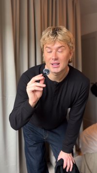 Black wireless microphone with a fuzzy windscreen, held in a hand. The background features soft, neutral-colored drapes.