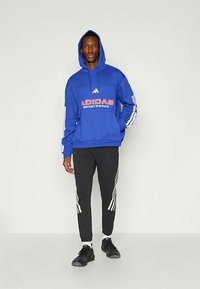 Μπλε φούτερ με κουκούλα που έχει γραφικό "ADIDAS SPORTWEAR", σε συνδυασμό με μαύρο παντελόνι με λευκές ρίγες στα πλάγια. Μαύρα αθλητικά παπούτσια ολοκληρώνουν την εμφάνιση.