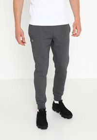 Grå bomullsjoggers med åtsittande ankle cuffs, som har en liten grön logotyp på övre vänstra sidan, tillsammans med svarta sportskor.