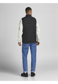 Gilet imbottito nero con cuciture orizzontali, indossato sopra una maglietta a maniche lunghe di colore chiaro, abbinato a jeans blu e sneaker nere.