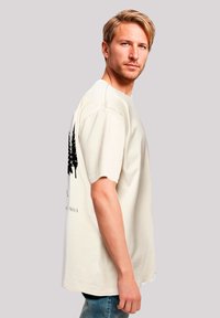 Beige cotton t-shirt with short sleeves, featuring a black tree graphic on the back. Model wearing distressed denim jeans.