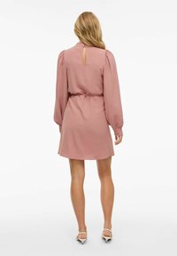 Rosa, texturerad klänning med långa puffärmar, cinchad midja och nyckelhålsdetalj i ryggen. Kjolslutningen når halvvägs upp på låret. Matchas med ljusa skor.