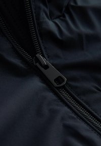 Fermeture éclair noire partiellement fermée sur un tissu sombre avec des coutures visibles et une texture lisse légèrement froissée.