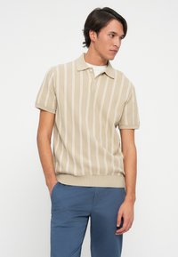 Beige knitted polo shirt with white vertical stripes, short sleeves, a collared neckline, and ribbed hem. Styled with blue pants.